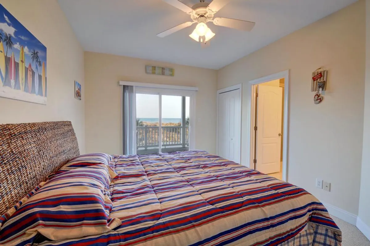 This interior image shows a cheerful bedroom with striped bedding, a large window offering a glimpse of the outdoors, and surfboard-themed artwork on the wall. The natural light streaming in hints at proximity to the beach, inviting thoughts of easy access to outdoor adventures.