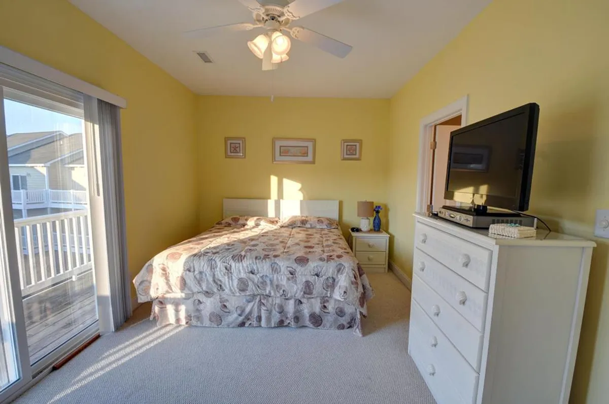 A cozy bedroom with patterned bedding, a television set atop a dresser, and a sliding glass door opening to a balcony. The space is filled with sunlight, creating a sense of openness and tranquility.