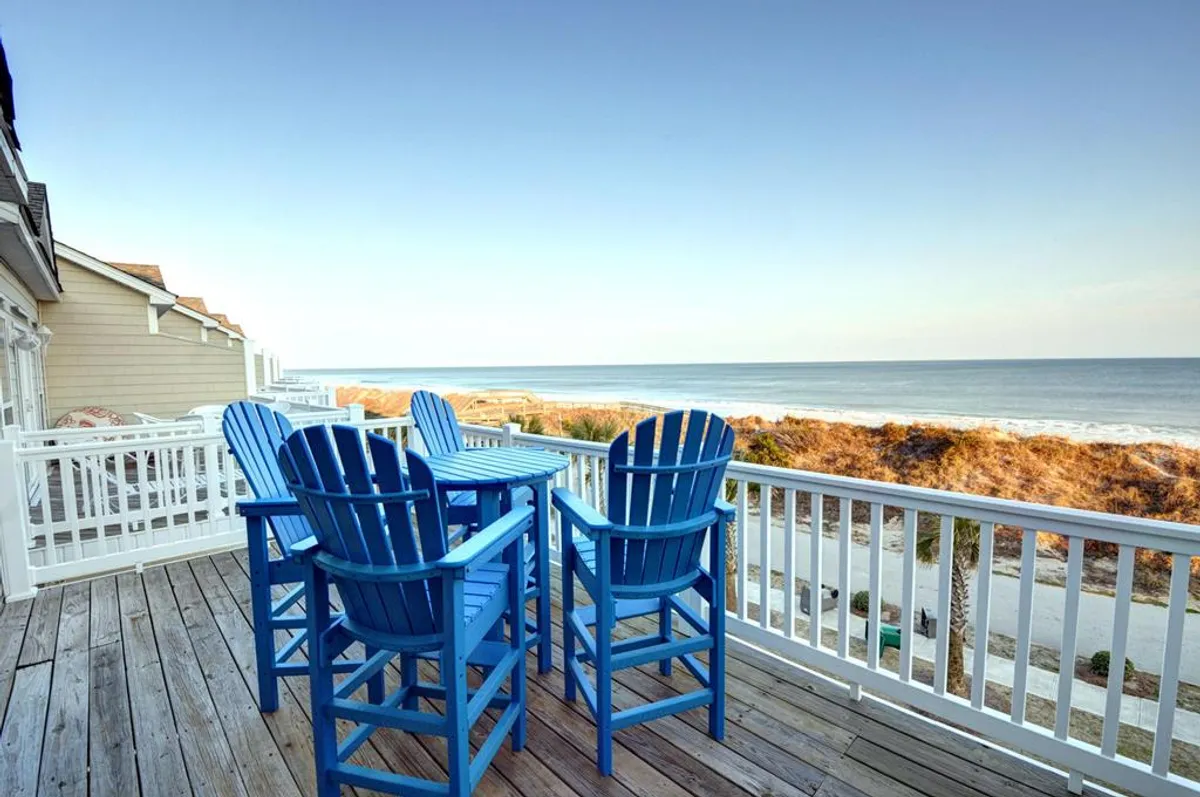 The image showcases a spacious wooden deck overlooking the ocean, featuring inviting blue Adirondack chairs perfectly positioned for relaxing, socializing, or soaking up breathtaking sunrise views. The peaceful ambiance and direct sightline to the waves create an irresistible outdoor living space ideal for vacation lounging.
