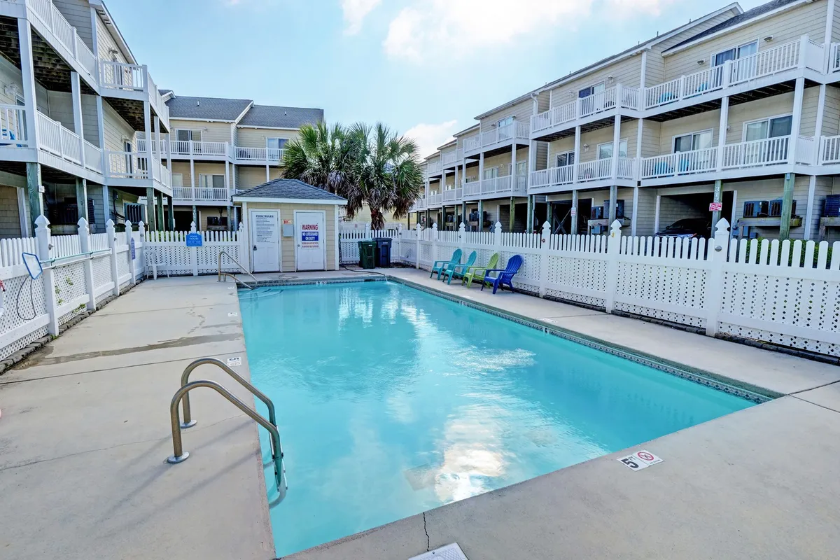 This image shows a lively swimming pool surrounded by palm trees, colorful lounge chairs, and a white picket fence. The atmosphere is cheerful and relaxed, with the inviting blue water glistening under the sunlight, suggesting an ideal setting for poolside relaxation and socializing with friends or family.