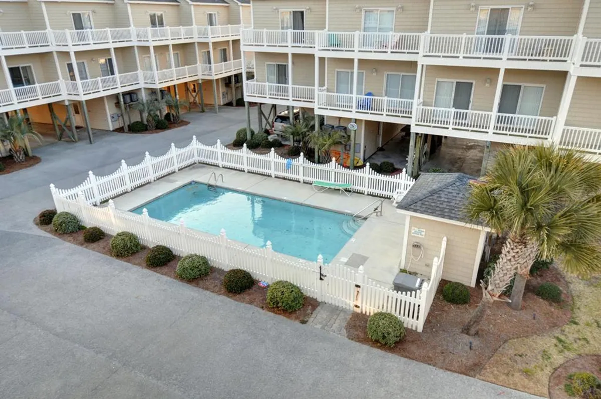 This exterior shot features the swimming pool framed by a white fence, with surrounding greenery and apartment balconies in the background. People can imagine swimming, sunbathing, or socializing by the pool, capturing the communal and inviting spirit.